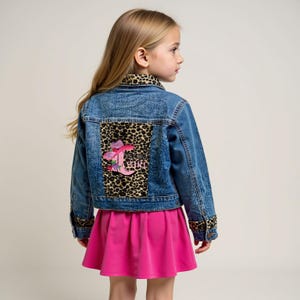 May include: A denim jacket with leopard print accents and a pink skirt. The jacket features a leopard print panel on the back with a pink embroidered design that says "GIRL". The jacket also has leopard print on the collar and cuffs.