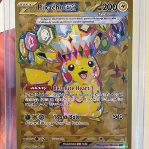 May include: A gold-colored Pikachu ex Tera trading card in a clear protective sleeve. The card features a crystalline Pikachu illustration, 200 HP, the Resolute Heart ability, and the Topaz Bolt attack. Text includes card details and game rules.
