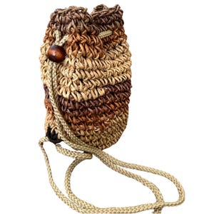 Puede incluir: Una bolsa con cordón tejido en tonos marrón, beige y bronceado. La bolsa tiene forma redondeada con cierre de cordón trenzado y un adorno de cuentas de madera. La textura de la bolsa es visible y el cordón es de color beige claro.