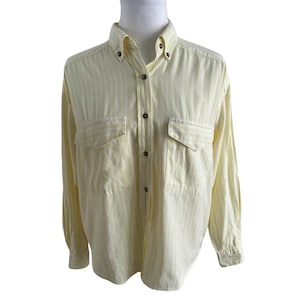 Könnte beinhalten: Ein Button-Down-Hemd mit gelb-weißem Streifenmuster. Das Hemd hat zwei Vordertaschen, lange Ärmel und einen Button-Down-Kragen. Das Hemd wird auf einer Schaufensterpuppe präsentiert.