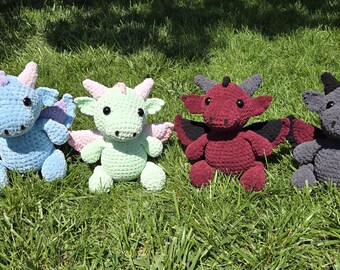 Puede incluir: Cuatro peluches de dragón de ganchillo en varios colores: azul claro con alas moradas, verde claro con alas rosas, burdeos con alas negras y gris con alas rojas. Cada dragón tiene cuernos y está sentado sobre hierba verde.