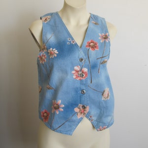 May include: A light blue denim vest adorned with a floral pattern featuring pink and peach-colored flowers. The vest has a V-neck, a button-up front, and a tailored fit. The floral print adds a touch of vintage charm to this denim vest.