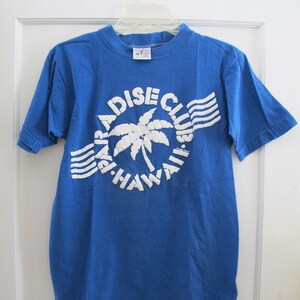 May include: A blue t-shirt with a white graphic featuring a palm tree and the words "Paradise Club" and "Hawaii". The shirt has short sleeves and a crew neck. The shirt is hanging on a white hanger.