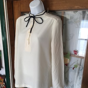 May include: Cream-colored long-sleeve blouse with a mandarin collar and a navy blue tie. The blouse has a button-down placket and is displayed on a white mannequin. The fabric appears to be lightweight and slightly textured.