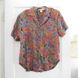 May include: A short-sleeved button-down shirt with a vibrant, abstract pattern in shades of pink, orange, blue, green, and black. The shirt has a collared neckline and a single chest pocket. It is hanging on a clear plastic hanger.