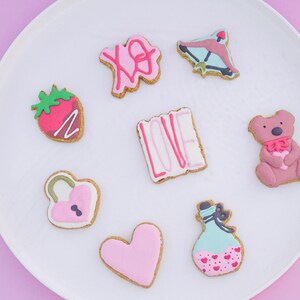 Puede incluir: Un plato blanco exhibe una colección de galletas de San Valentín. Las galletas están decoradas con glaseado rosa y rojo en varias formas, incluyendo una fresa, un corazón, un oso de peluche y la palabra "LOVE".