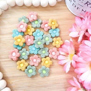 以下が含まれることがあります： ピンク、黄色、青、緑の色合いの、花型のカラフルなお菓子。明るい茶色の木製の表面に配置され、白いビーズの列とピンクの花が背景にあります。