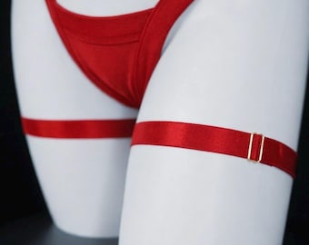 May include: A pair of red elastic leg garters featuring a smooth, satin-like finish and a small, rectangular gold-toned metal slider for adjustment. The garters are positioned horizontally around the upper thigh area, providing a simple design.