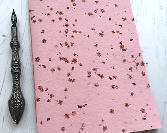 May include: A pink notebook with a floral pattern made from pressed flowers. The notebook has a red spine and is closed.