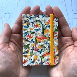 May include: A small, rectangular notebook with a colorful floral pattern. The design features red, yellow, blue, and green leaves and flowers on a white background. A yellow elastic band secures the notebook. The notebook is held in open hands.