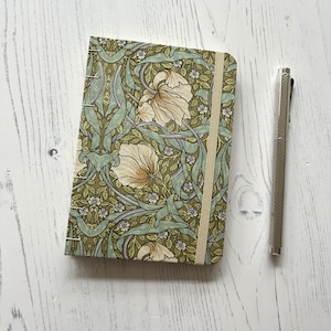 May include: A small, green and white floral patterned notebook with a cream ribbon bookmark. The notebook is closed and sitting on a white wooden surface.