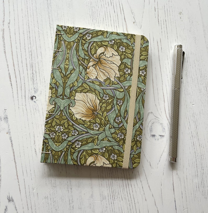 May include: A small, green and white floral patterned notebook with a cream ribbon bookmark. The notebook is closed and sitting on a white wooden surface.