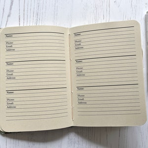 May include: A small, brown, lined notebook with a silver pen. The notebook has blank pages for writing names, phone numbers, emails, and addresses.