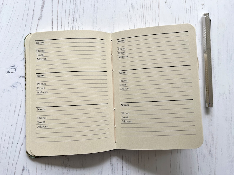 May include: A small, brown, lined notebook with a silver pen. The notebook has blank pages for writing names, phone numbers, emails, and addresses.