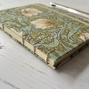 May include: A green and white floral patterned notebook with a spine bound with white thread. The notebook is closed and lying on a white wooden surface.