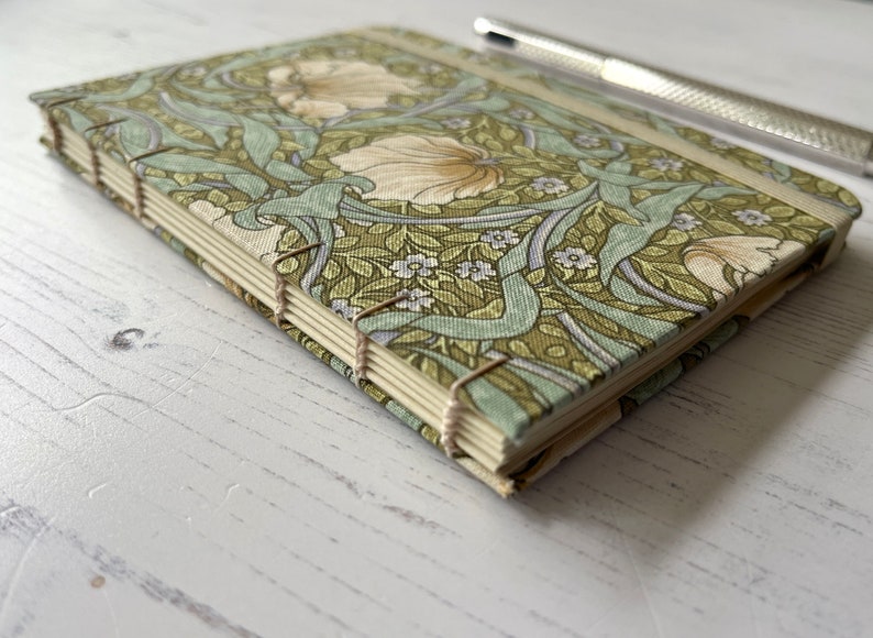 May include: A green and white floral patterned notebook with a spine bound with white thread. The notebook is closed and lying on a white wooden surface.