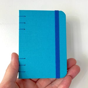 May include: A small, turquoise notebook with a blue elastic band closure and stitched binding. The notebook is held in a hand, showcasing its compact size and textured cover. The notebook's dimensions are approximately 9 cm by 13 cm.