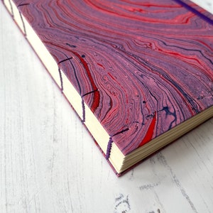 May include: A purple and red marbled journal with a purple ribbon bookmark. The journal is closed and lying on a white wooden surface.