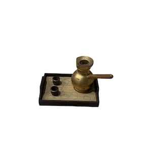 Puede incluir: Un pequeño juego de café dorado sobre una bandeja rectangular. El juego incluye una cafetera pequeña con un mango largo y dos tazas pequeñas. La bandeja tiene un diseño decorativo.