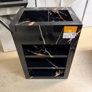 May include: A black marble vanity with gold veining, featuring an open top and two shelves. The vanity has a rectangular shape and is displayed in a showroom setting. The surface has a glossy finish.
