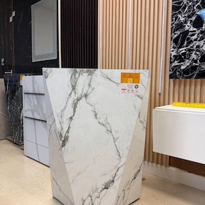 May include: A geometric, marble-patterned pedestal sink with a white and gray veined design. The sink has a multi-faceted shape and is displayed in a showroom setting. The sink is a modern design.