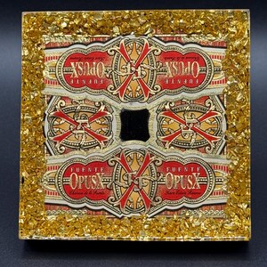 May include: Square resin art piece featuring Fuente Opus X cigar bands in red, gold, and cream. The bands are arranged around a central black square, encased in a gold glitter border. The artwork is set against a black background.