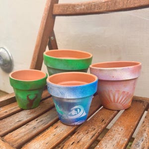 May include: Four small, hand-painted terracotta flower pots. The pots are painted in various colors, including green, blue, and pink, with decorative designs. The pots are sitting on a wooden chair.