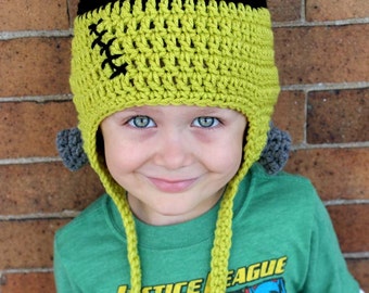 May include: A young child wearing a green and black crocheted hat with ear flaps and pom-poms. The hat has a stitched design on the front. The child is wearing a green shirt with the words "Justice League" printed on it.