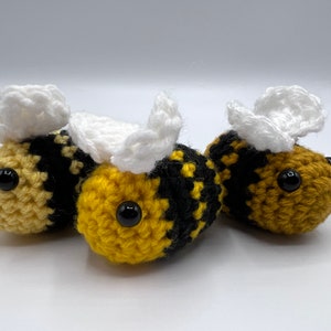 May include: Three crocheted bumblebees with black and yellow striped bodies and white wings. The bumblebees have black button eyes.