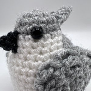 May include: A crocheted gray and white bird with black eyes. The bird is made with a textured yarn and has a fluffy appearance.