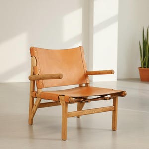 May include: A light brown leather and wood chair with armrests. The chair has a light brown leather seat and back, and the frame is made of light-colored wood. The chair has a simple, minimalist design and is suitable for indoor use.