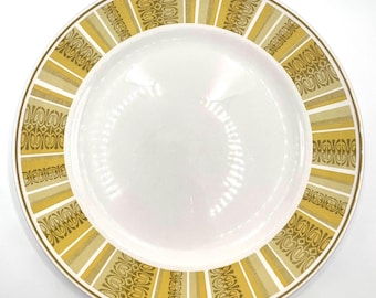 Puede incluir: Un plato de cerámica blanco con un borde decorativo. El borde presenta un patrón repetitivo de formas geométricas y rayas verticales de color verde oliva y dorado. El plato tiene un borde ancho y una zona central rebajada.