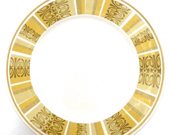 Puede incluir: Un plato blanco con un borde decorativo. El borde presenta un patrón repetitivo de formas geométricas doradas y beige y rayas verticales. El plato es redondo y tiene un diseño simple y elegante.