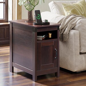 May include: A dark brown wooden side table with a charging station on top. The table has a small cabinet with a door and a shelf. A smartphone is on the charging station, showing a charging symbol. The table is next to a beige sofa.