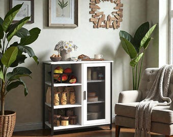 May include: A white and black cabinet with open shelves and a glass-fronted door. The cabinet is filled with various items, including food and decorative objects. A potted plant and framed artwork are visible in the background, creating a cozy interior setting.