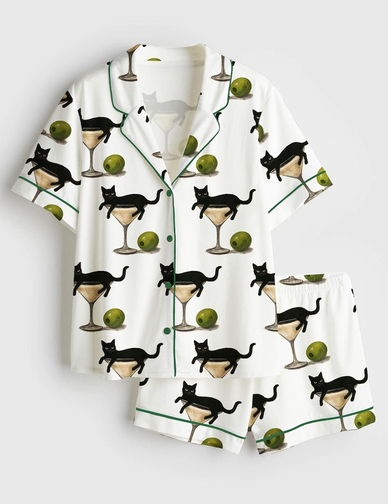 May include: White pyjama set featuring a pattern of black cats in martini glasses and green olives. The short-sleeved top and shorts have green trim.
