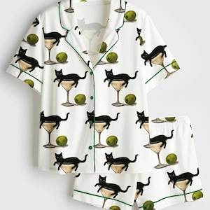 May include: White pajama set featuring a pattern of black cats in martini glasses and olives. The short-sleeved top and shorts have green trim. The set is made of a soft, lightweight fabric.