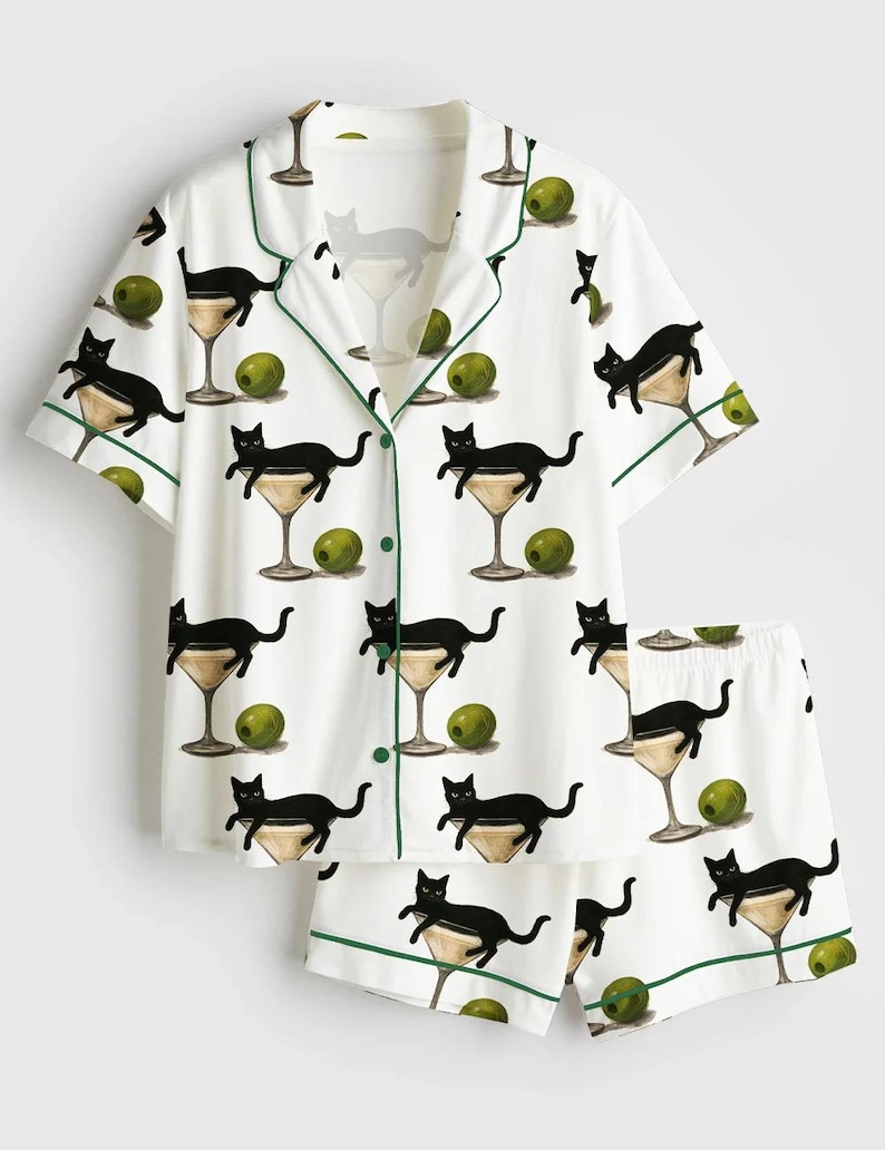 May include: White pajama set featuring a pattern of black cats in martini glasses and olives. The short-sleeved top and shorts have green trim. The set is made of a soft, lightweight fabric.