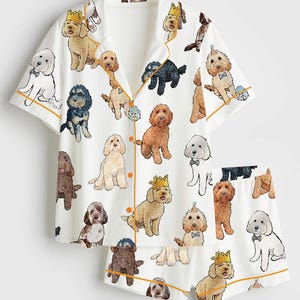 May include: White pajama set with a repeating pattern of illustrated dogs in various colors and poses. The set includes a short-sleeved button-up shirt and matching shorts, both trimmed with orange piping.