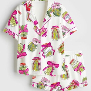 Puede incluir: Conjunto de pijama blanco con un estampado de tarros de pepinillos. La camiseta de manga corta y los pantalones cortos presentan un dise&ntilde;o repetido de tarros de pepinillos con lazos rosas y la palabra "Pickles". El ribete es rosa.