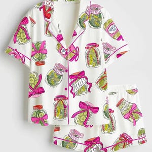Puede incluir: Conjunto de pijama blanco con un estampado de tarros de pepinillos. La parte superior de manga corta y los pantalones cortos presentan un dise&ntilde;o repetido de tarros de pepinillos con lazos rosas y la palabra "Pickles". El conjunto tiene ribetes rosas.