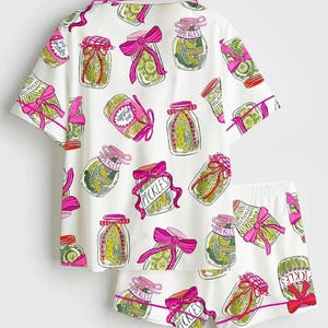 Puede incluir: Conjunto de pijama blanco con un estampado de tarros de pepinillos. La camiseta de manga corta y los pantalones cortos presentan ilustraciones de tarros de pepinillos con lazos rosas y la palabra "Pickles". El ribete es rosa.