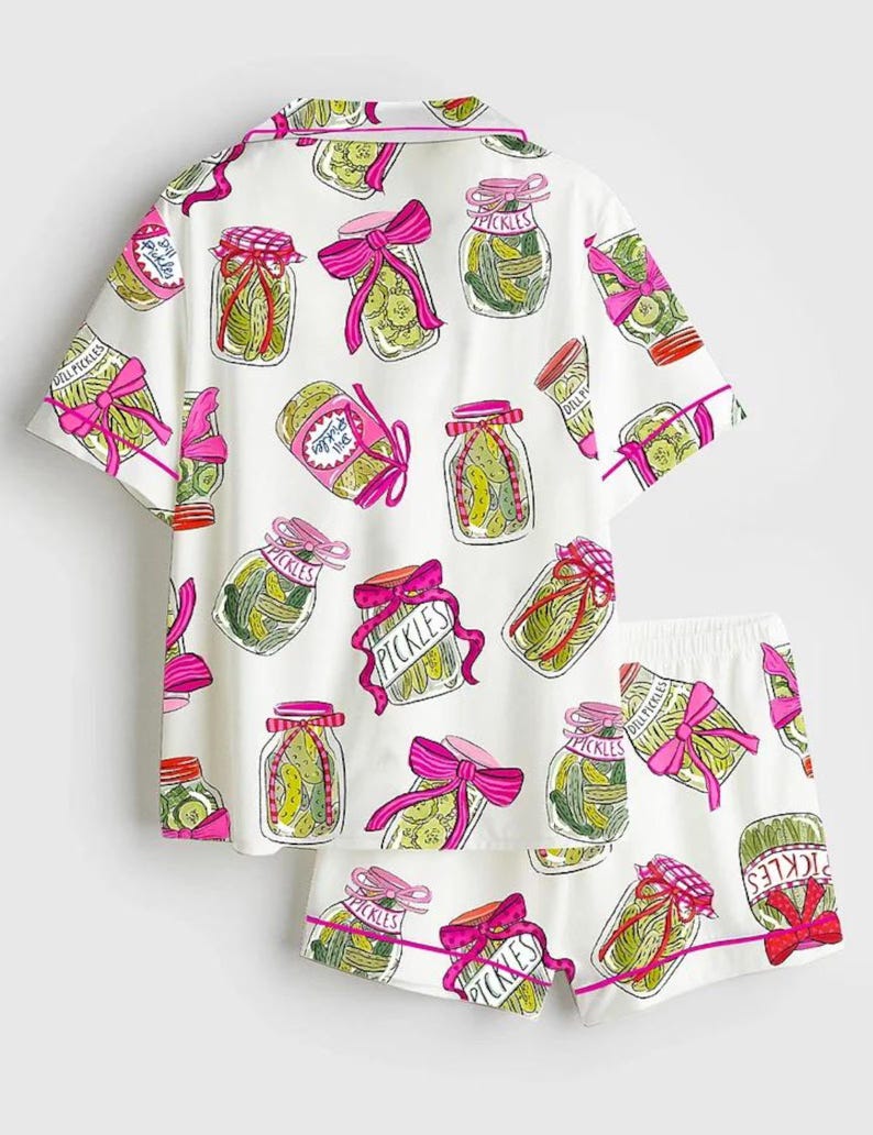 Puede incluir: Conjunto de pijama blanco con un estampado de tarros de pepinillos. La camiseta de manga corta y los pantalones cortos presentan ilustraciones de tarros de pepinillos con lazos rosas y la palabra "Pickles". El ribete es rosa.