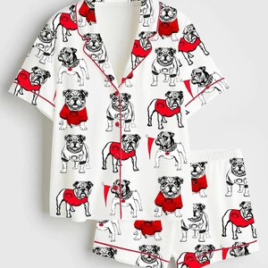 May include: White pyjama set featuring a repeating pattern of bulldog illustrations in black and white, some wearing red sweaters. The set includes a button-down short-sleeved top and matching shorts, both trimmed with red piping.