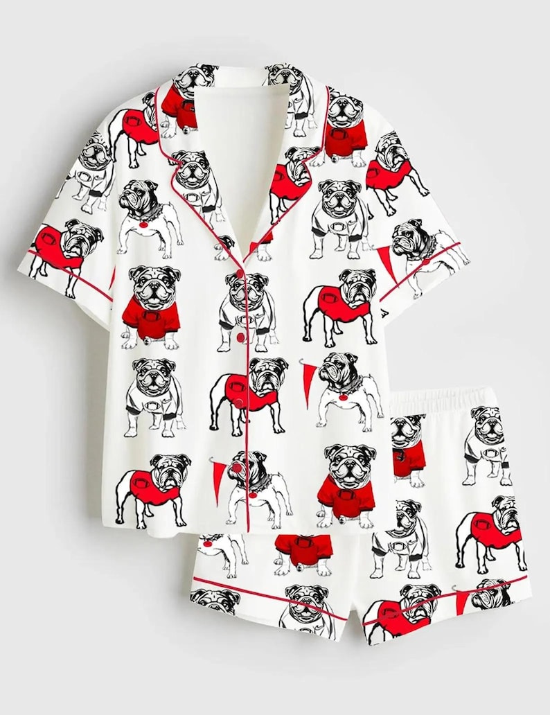 May include: White pyjama set featuring a repeating pattern of bulldog illustrations in black and white, some wearing red sweaters. The set includes a button-down short-sleeved top and matching shorts, both trimmed with red piping.
