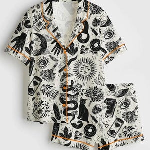 May include: A two-piece pajama set featuring a white background with black occult-themed illustrations. The short-sleeved top and shorts are trimmed with orange piping. The design includes moons, eyes, hands, snakes, cats, and crystals.