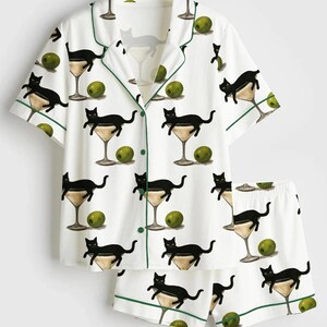 May include: White pyjama set featuring a pattern of black cats in martini glasses with olives. The short-sleeved top and shorts have green trim. The button-down top has a notched collar.
