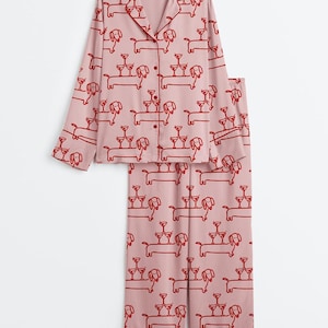 K&ouml;nnte beinhalten: Zweiteiliger Schlafanzug in Hellrosa mit einem sich wiederholenden Muster aus roten Dackeln, die gestapelte Martinigl&auml;ser tragen. Das Set besteht aus einem Oberteil mit Kn&ouml;pfen und langen &Auml;rmeln sowie einer passenden Hose.