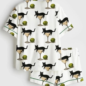 May include: White pyjama set with a repeating pattern of black cats in martini glasses and green olives. The short-sleeved top and shorts have green trim.