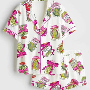 Puede incluir: Conjunto de pijama blanco con un estampado de tarros de pepinillos. La camiseta de manga corta y los pantalones cortos presentan un dise&ntilde;o repetido de tarros de pepinillos con lazos rosas y la palabra "Pickles". El conjunto tiene ribetes rosas.
